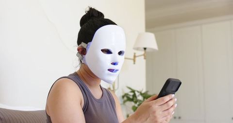 Home led facial mask: woman using light therapy while checking smartphone