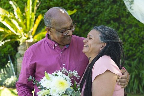 Senior couple celebrating anniversary in sunlit garden holding white bouquet, laughing