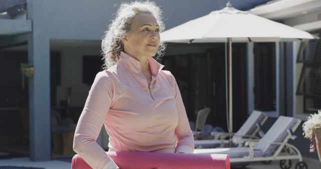 This image shows a senior woman holding a yoga mat outdoors on a sunny day, indicating an active and healthy lifestyle. Ideal for use in articles, advertisements, or blogs promoting senior fitness, wellness, outdoor activities, and healthy living among retirees.
