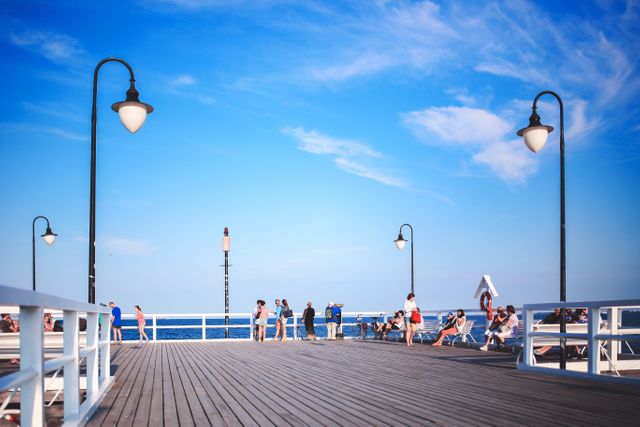 People enjoying a sunny day on a scenic ocean pier under a clear blue sky. This setting is perfect for themes related to vacations, outdoor relaxation, leisurely strolls, tourism, and travel destinations. The lampposts lining the boardwalk add charm to the scene and convey a sense of a peaceful getaway by the water.