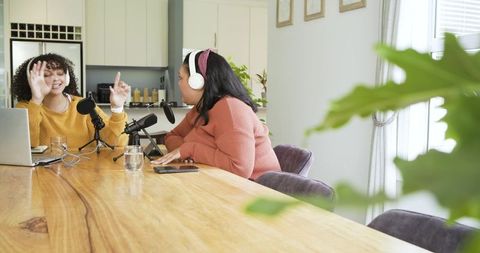 Diverse women cohost duo recording cozy home kitchen podcast with microphones, laptop, headphones
