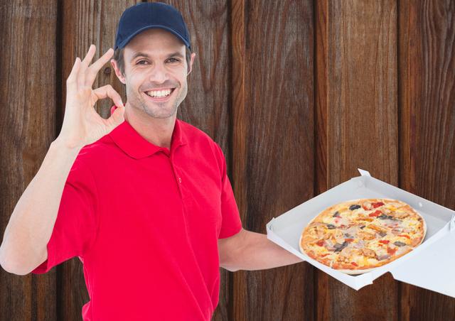 This image shows a cheerful deliveryman in a red uniform and cap holding a pizza box and making an OK gesture. The wooden background adds a rustic feel. Ideal for use in advertisements for food delivery services, fast food promotions, customer service training materials, and restaurant marketing campaigns.