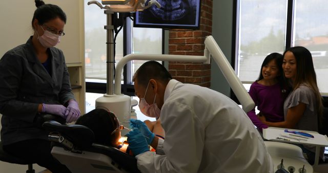 Dentist providing treatment to young boy in dental clinic while mother and sister watch. Dental assistant preparing tools. Depicts environment of pediatric dentistry. Suitable for use in health, medical practice, family care advertisements.
