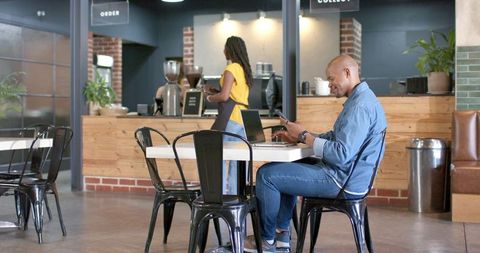 African american man working on laptop and smartphone in modern industrial coffee shop