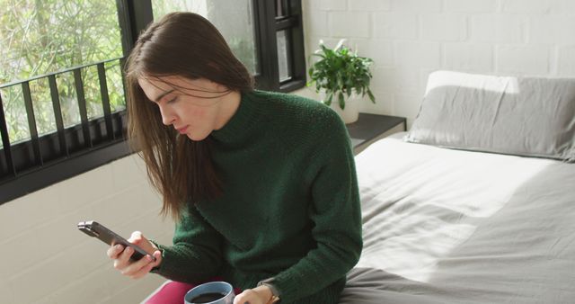 Caucasian non-binary transgender woman sitting on bed and using smartphone. spending quality time at home alone, body inclusivity.