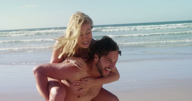 A young Caucasian couple enjoys a playful moment on a sunny beach, with the woman piggybacking on the man, both smiling joyfully. Their carefree fun captures the essence of a romantic getaway by the sea.