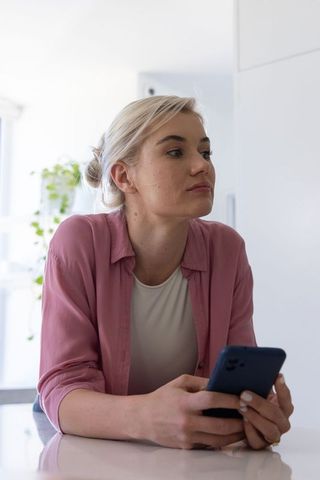 Contemplative woman holding smartphone at modern kitchen counter, minimalist lifestyle
