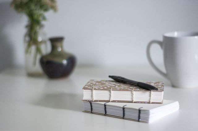 Notebooks with a pen placed on a minimalist desk. A decorative vase with flowers and a white coffee mug are also in the background. This can be used for themes related to creative writing, office work, study environment, productivity, and desk organization.