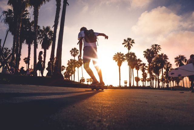 Teenager skateboarding along a California beach path lined with palm trees against a dramatic sunset sky. Ideal for themes related to summer, outdoor recreation, travel, youth culture, and coastal lifestyles.