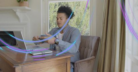 Mid-adult black man reaching for smartphone at laptop in workspace with neon light streaks