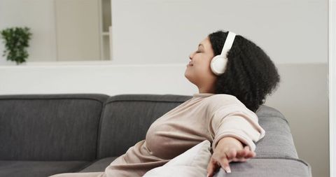 Young woman reclining on grey sofa wearing white headphones in minimalist room