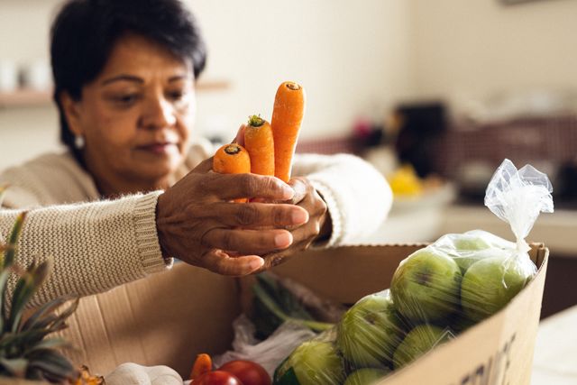 Biracial mature woman removing carrots from cardboard box while unpacking groceries in kitchen. Home, food, shopping, unaltered, lifestyle and retirement concept.