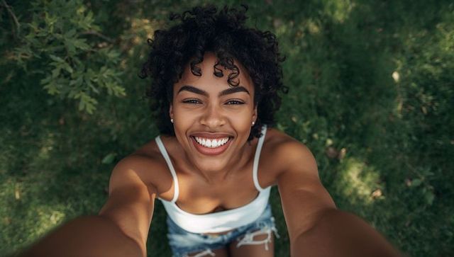Smiling woman taking overhead selfie on sunlit grass, casual summer outfit with denim shorts