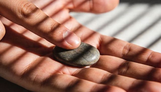 Holding smooth grey pebble in open palm under sunlight with blind shadow stripes