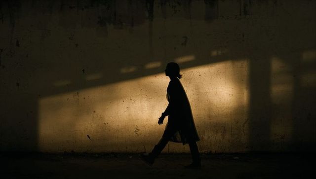 Silhouetted figure walking in long coat through slanted light on grungy tunnel wall