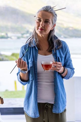 Senior woman painter on lakeside terrace holding teacup and brush, coastal view, serene