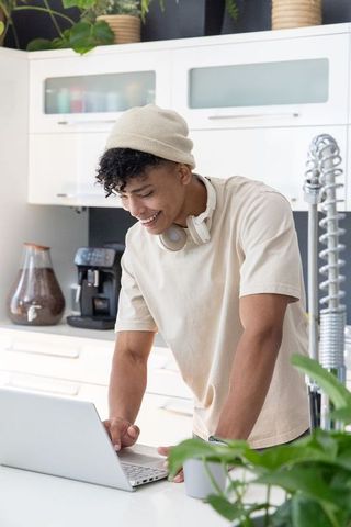 African american man leaning over laptop in kitchen smiling with beanie and headphones