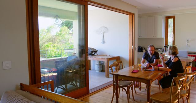 Couple conversing at home dining table, with a view of greenery from the balcony. Ideal for concepts of home life, couple interactions, casual conversations, relaxed atmospheres, and modern homes. Useful for home decor, furniture, relationship topics, and family lifestyle themes.