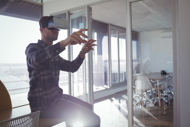 Businessman using VR headset in a modern office, gesturing as he interacts with virtual elements. Ideal for illustrating concepts of innovation, technology in business, and modern workplace environments. Suitable for use in articles, presentations, and marketing materials related to virtual reality, tech advancements, and creative business solutions.