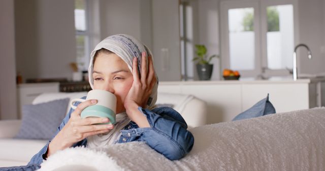 This image captures a woman in a hijab relaxing on a couch with a cup of coffee in a cozy home environment. The background features an open kitchen, emphasizing a comfortable and serene atmosphere. This image can be used in content related to relaxation, home comfort, cultural representation, lifestyle blogs, or advertisements focusing on home and personal well-being.