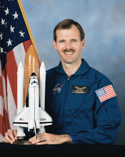 Astronaut Steven L. Smith wearing a blue NASA flight suit, standing next to a detailed model of the Space Shuttle. The American flag is prominently displayed in the background, symbolizing national pride and space achievements. Great for educational materials about space, NASA programs, inspirational content about space exploration, and promoting STEM education.