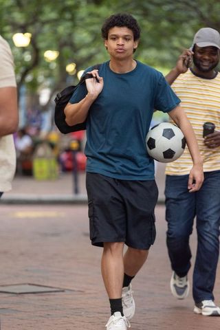 Young adult walking in city plaza with soccer ball and pack while friend talks on phone