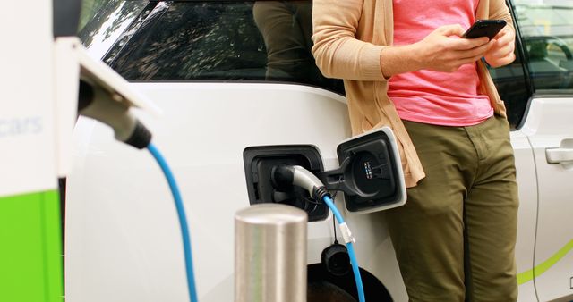 A person is charging an electric vehicle at a charging station, with copy space. They are focused on their smartphone while the car is being powered up, highlighting the convenience of modern electric transportation.