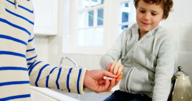 Child and parent sitting in kitchen, sharing a carrot. Use in parenting, family bonding, healthy eating, and lifestyle content.