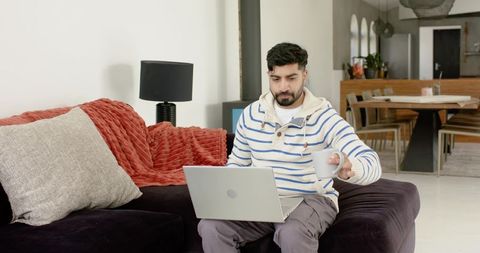 Indian man working from home on laptop holding coffee mug on cozy modern sofa in open-plan room