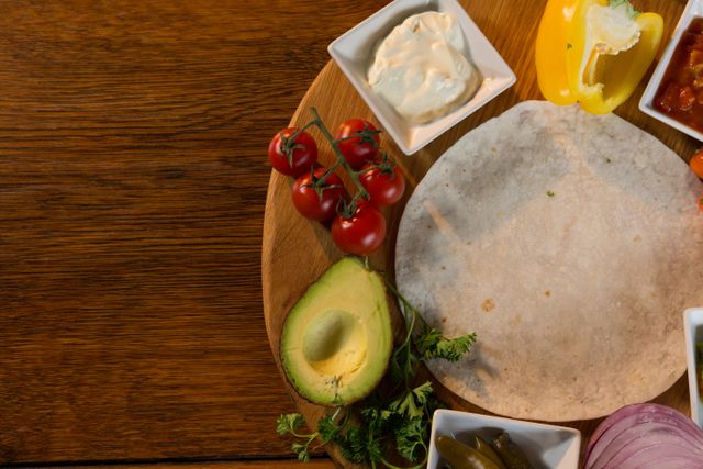 Mexican food ingredients arranged on a wooden table, including tortilla, avocado, cherry tomatoes, bell pepper, salsa, and sour cream. Ideal for use in culinary blogs, recipe websites, cooking tutorials, and healthy eating promotions.