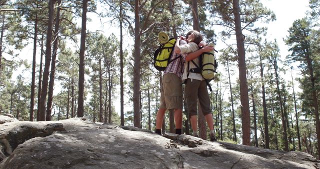 A middle-aged Caucasian couple wearing backpacks share a kiss on a rocky outcrop in a forest, with copy space. Their embrace amidst the serene natural landscape suggests a moment of connection and adventure during a hiking trip.