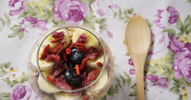 A glass of yogurt parfait topped with banana slices, blueberries, and goji berries sits next to a wooden spoon on a floral tablecloth. The parfait offers a healthy snack option, beautifully presented and ready to enjoy.