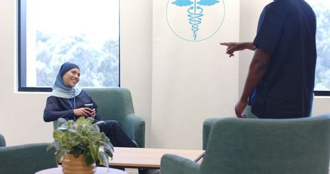 Muslim nurse holding coffee mug smiling and chatting with coworker in staff lounge