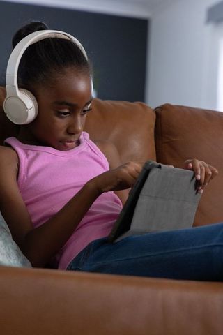 African girl relaxing on brown sofa listening with headphones and using tablet for learning