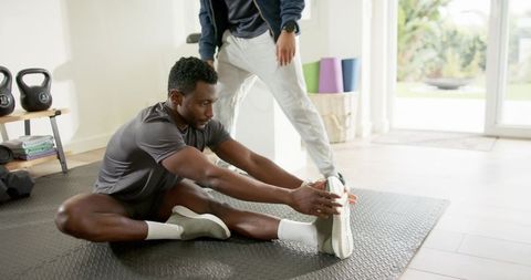 African american man stretching with male trainer on home workout mat near kettlebells