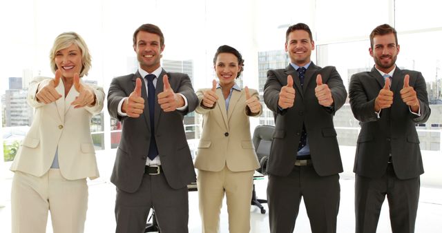 Group of diverse business professionals standing in a modern office, all giving thumbs up and smiling. Suitable for themes about teamwork, corporate success, positive work culture, and business achievements. Can be used for corporate websites, presentations, promotional materials, and professional blogs.