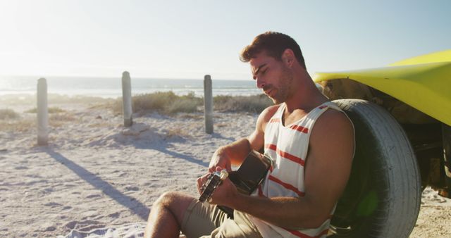 Young man in a striped tank top sitting on the sandy beach playing guitar during a sunset. Ideal for themes related to music, relaxation, summer vacations, outdoor leisure, and peaceful moments.