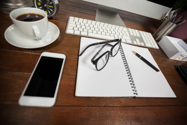 This image depicts a neatly organized workspace featuring a cup of coffee, a notebook, a smartphone, glasses, and a pen on a wooden desk. Ideal for illustrating concepts related to productivity, remote work, office environments, and professional settings. Suitable for use in articles, blog posts, and marketing materials focused on business, technology, and work-life balance.