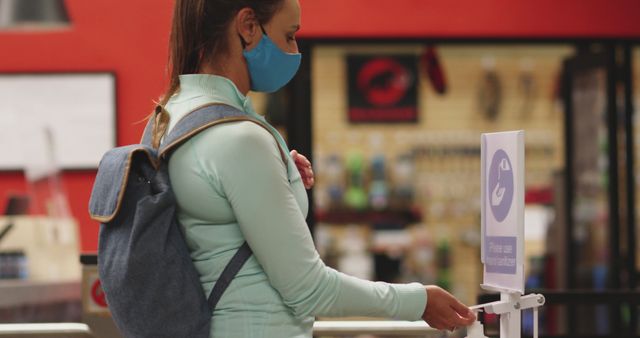 Scene showing a young woman using a hand sanitizer dispenser at the entrance of a store while wearing a mask. The image is useful for illustrating concepts related to personal hygiene, COVID-19 precautions, health safety measures, and responsible behavior in public spaces.