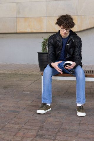 Teenage male wearing leather jacket and glasses sitting on bench holding vr headset in urban plaza