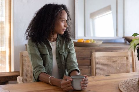 Woman holding mug and looking through window at wooden dining table in cozy rustic home