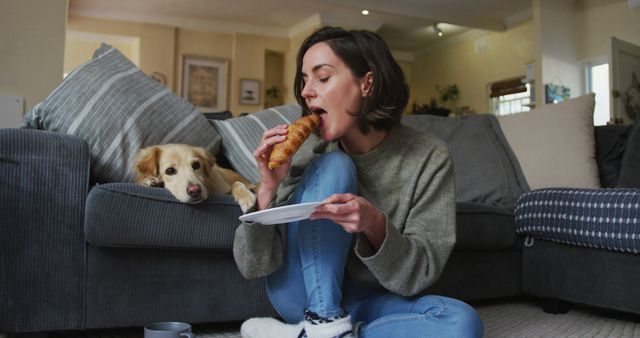 Smiling caucasian woman eating croissant and stroking her pet dog on sofa next to her. pet companionship, domestic life and working from home concept.