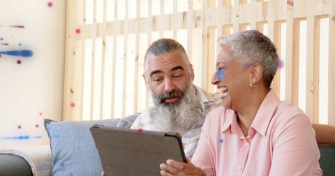 Mature couple laughing while sharing tablet on cozy sofa in sunlit wooden living room