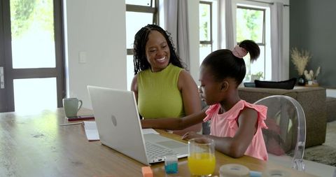 African american mother helping daughter homeschooling on laptop at sunlit wooden table