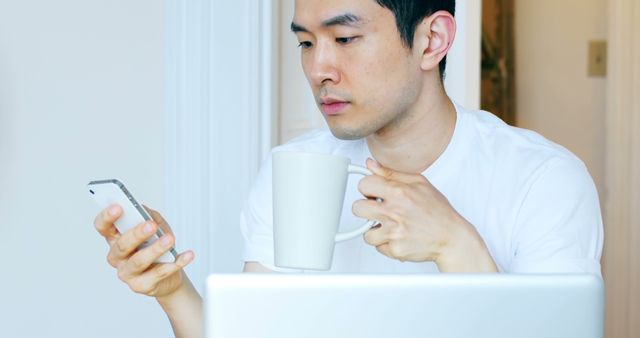Young man is holding a cup of coffee and looking at his smartphone while sitting at his home office with a laptop in front of him. He appears focused and concentrated, suggesting productivity and work-from-home scenarios. This image usable for home office, remote work, technology, multitasking themes and productivity tips.