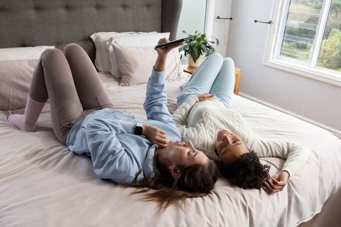 Two women lounging on bed holding tablet, wearing cozy loungewear, relaxed morning moment