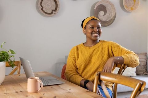 African american woman working from home with laptop cozy mustard sweater and headband