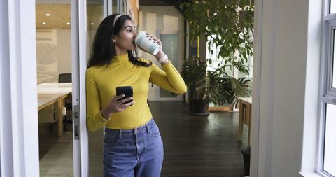 Indian professional drinking coffee and checking smartphone in modern office doorway