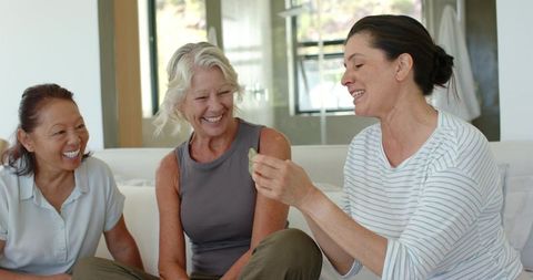 Laughing diverse women sitting on sofa sharing green leaf in bright modern living room