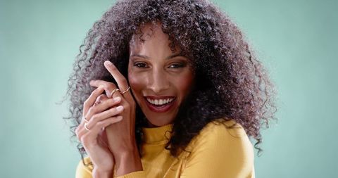 Smiling woman wearing mustard top showing thin rings and curly hair against mint backdrop